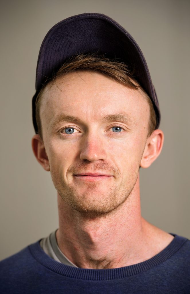 A photo of Fergus from the shoulder up, smiling at the camera. Fergus is wearing a baseball cap.