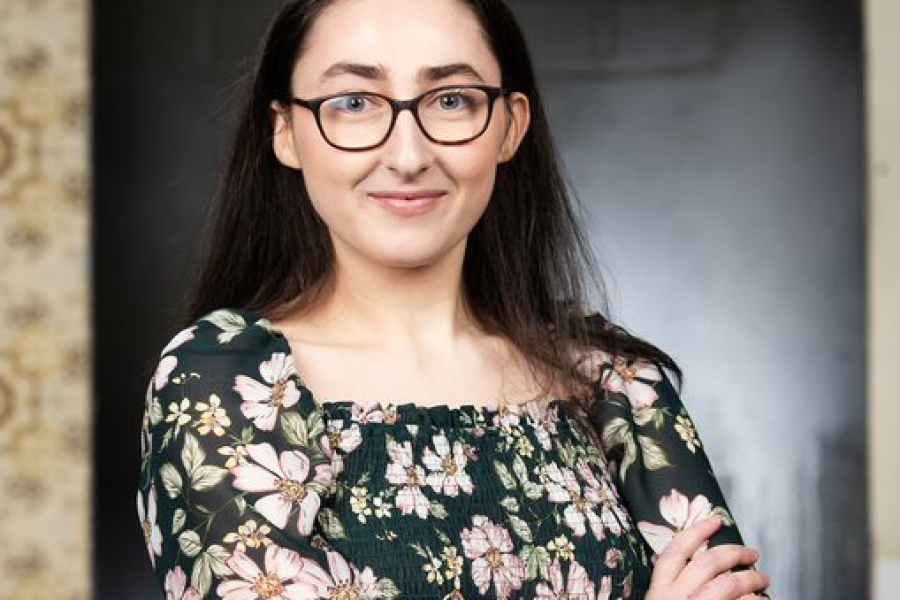 A photo of Joanna stood in a doorway with her arms folded, smiling at the camera. She is wearing a black top with a flower pattern and wears glasses.